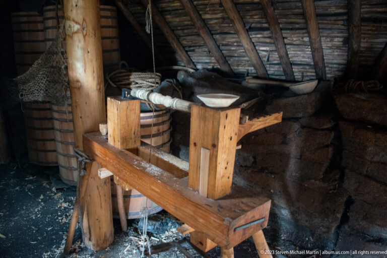 Heavy wooden textile-working frame with wound fiber and cordage inside a reconstructed turf-walled building.