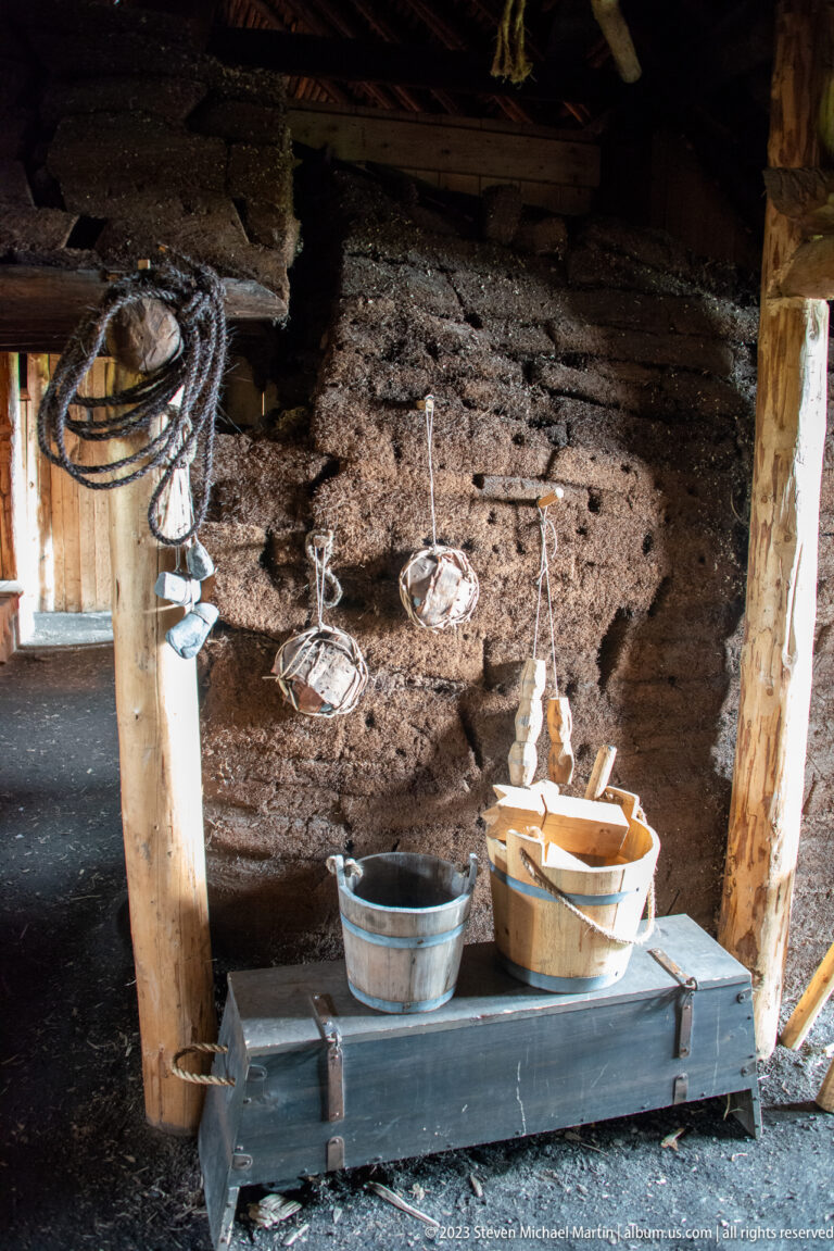 Hanging vessels suspended above wooden tubs and containers against a dark turf wall inside a reconstructed dwelling.