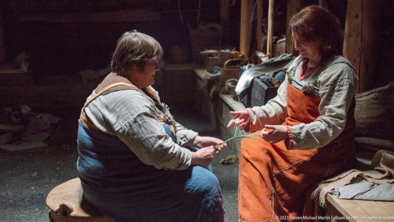 Two costumed interpreters seated indoors while handling bright green fiber or cord during a living-history demonstration.