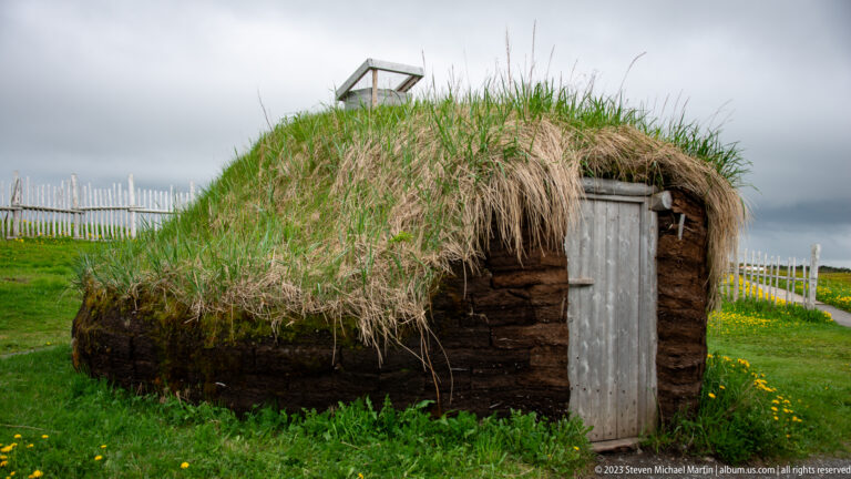 Small reconstructed turf-covered building with a simple plank door standing amid green grass and yellow wildflowers.