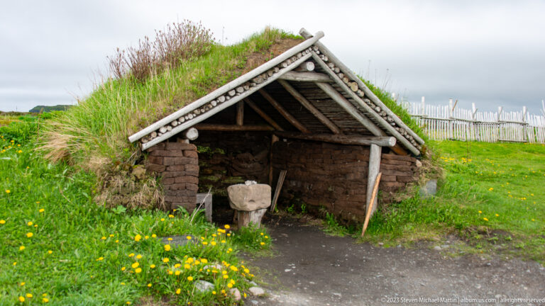 Open-fronted turf building with timber gable, wooden work fixture, meadow flowers, and palisade fence.