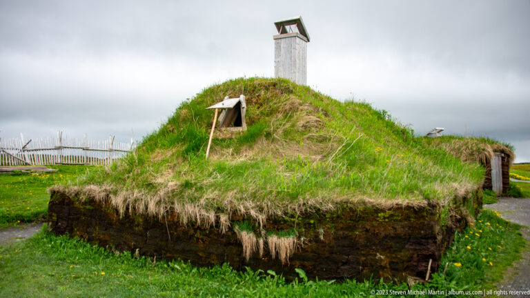 Low turf-covered reconstructed building with tall square roof vent and smaller hooded opening under gray sky.
