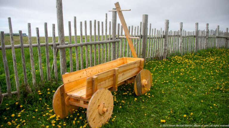 Wooden two-wheeled cart with solid disc wheels and long handle beside a tall palisade fence and yellow flowers.