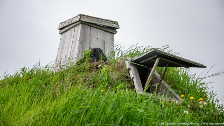 Square wooden roof vent and smaller hooded opening projecting from dense grass on a reconstructed turf roof.