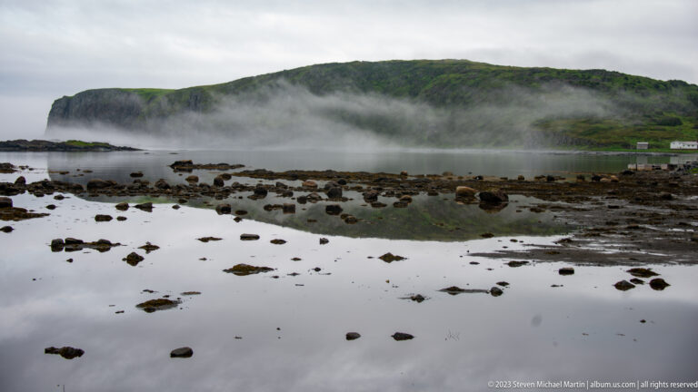 Shallow tidal water dotted with dark rocks beneath a long green headland wrapped in low fog.