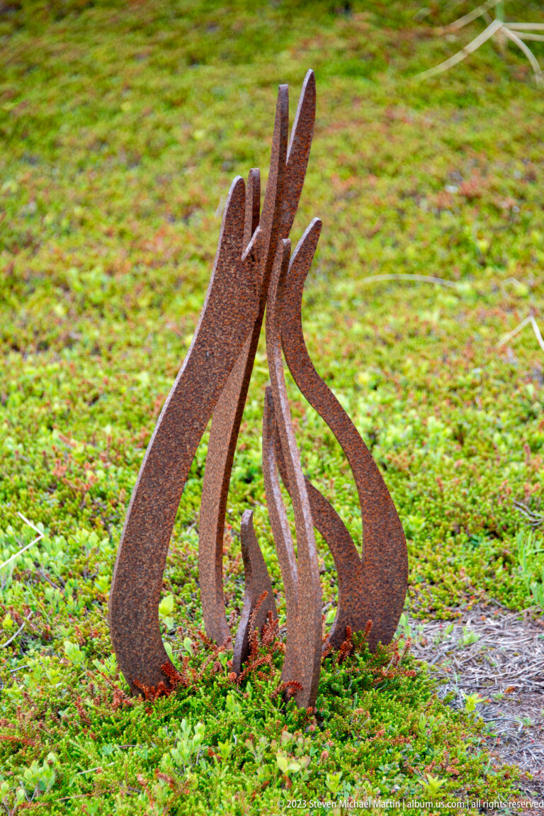 Small rusted-steel sculpture formed from curving strips shaped like rising flames on green groundcover.