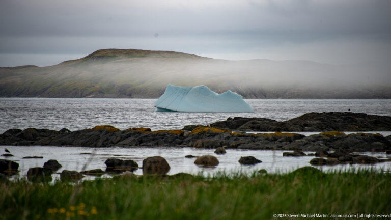 Blue-white iceberg offshore beyond dark tidal rocks, with a long fog-covered headland in the background.