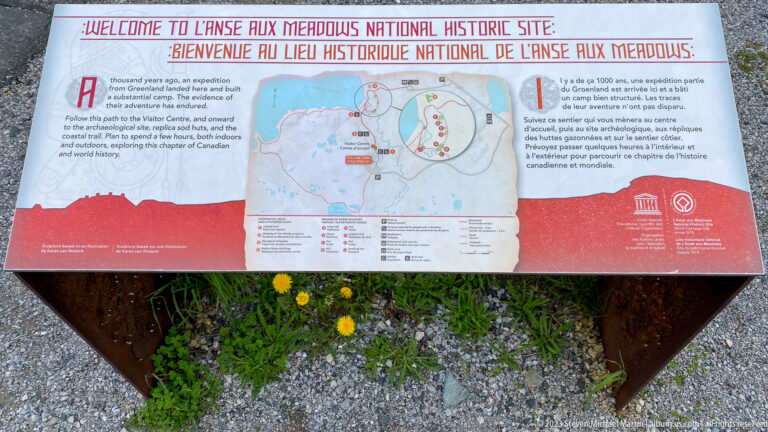 Bilingual outdoor welcome panel with map of L’Anse aux Meadows National Historic Site, mounted above gravel and flowers.