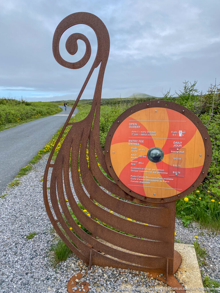Rusted steel Viking-ship-shaped sign supporting a circular orange orientation diagram beside a paved path.
