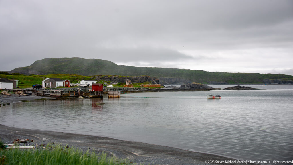 Small waterfront sheds and wharves beside a calm cove, with low green hills under an overcast sky.