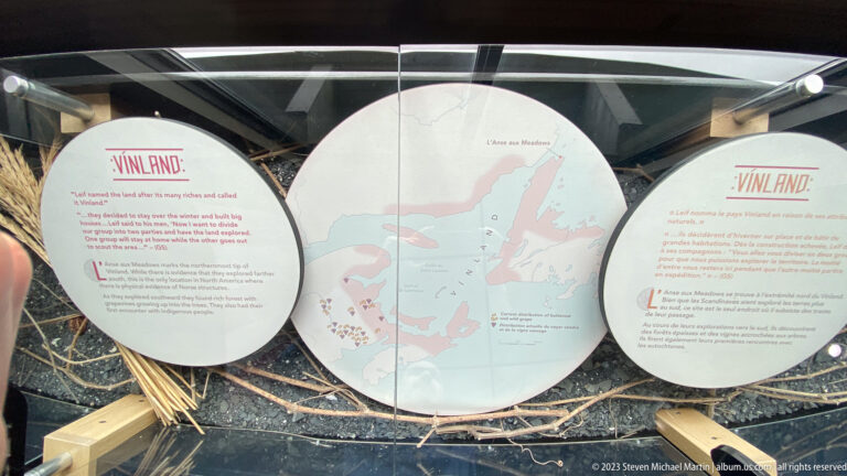 Large circular map of Newfoundland flanked by bilingual Vinland panels and plotted interpretive symbols.