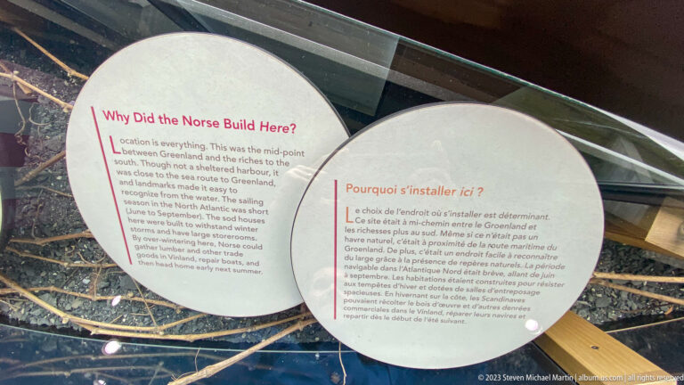 Bilingual Why Did the Norse Build Here interpretive panel displayed beneath glass with natural materials below.