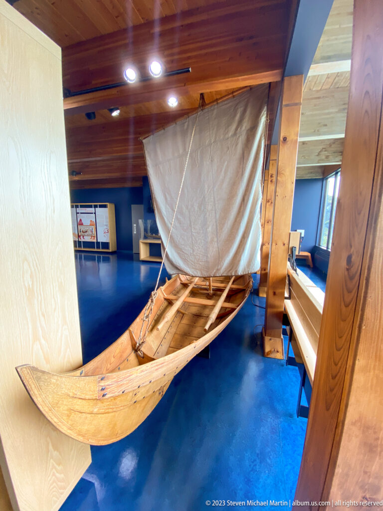 Full-scale wooden Norse-style boat with raised pale square sail displayed indoors in the visitor centre.