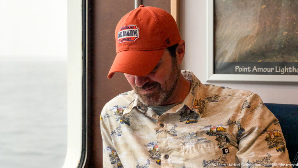 Man in an orange Expedition 51° Central cap seated indoors beneath a framed Point Amour Lighthouse print.