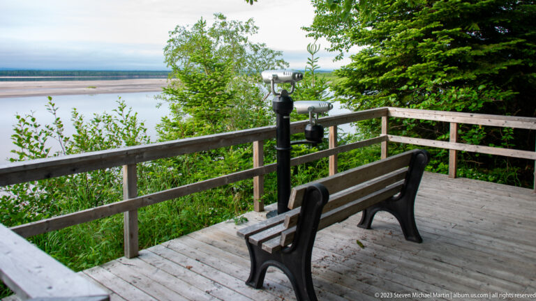 Wooden overlook deck with a bench and fixed viewing scope facing water and exposed flats through trees.