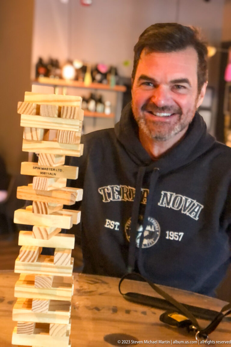 Close portrait of a smiling man beside a tall, uneven tower of stacked wooden blocks.