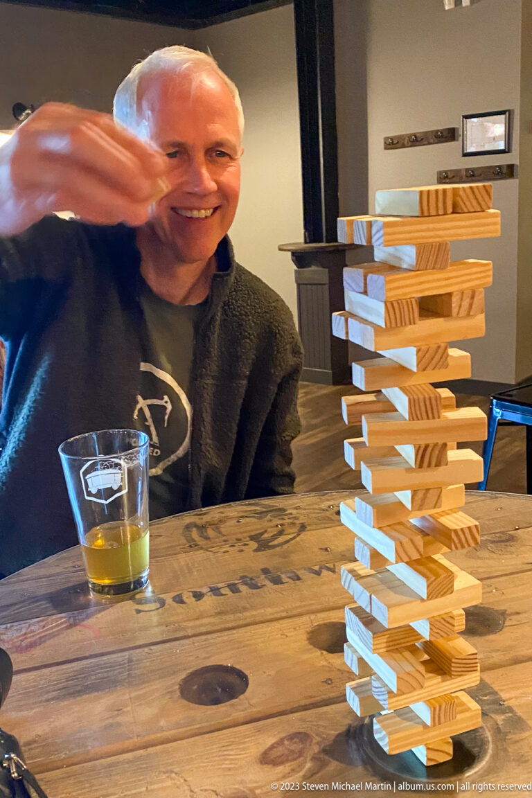 Seated smiling man beside a tall wooden block tower and a partly filled beer glass on the table.