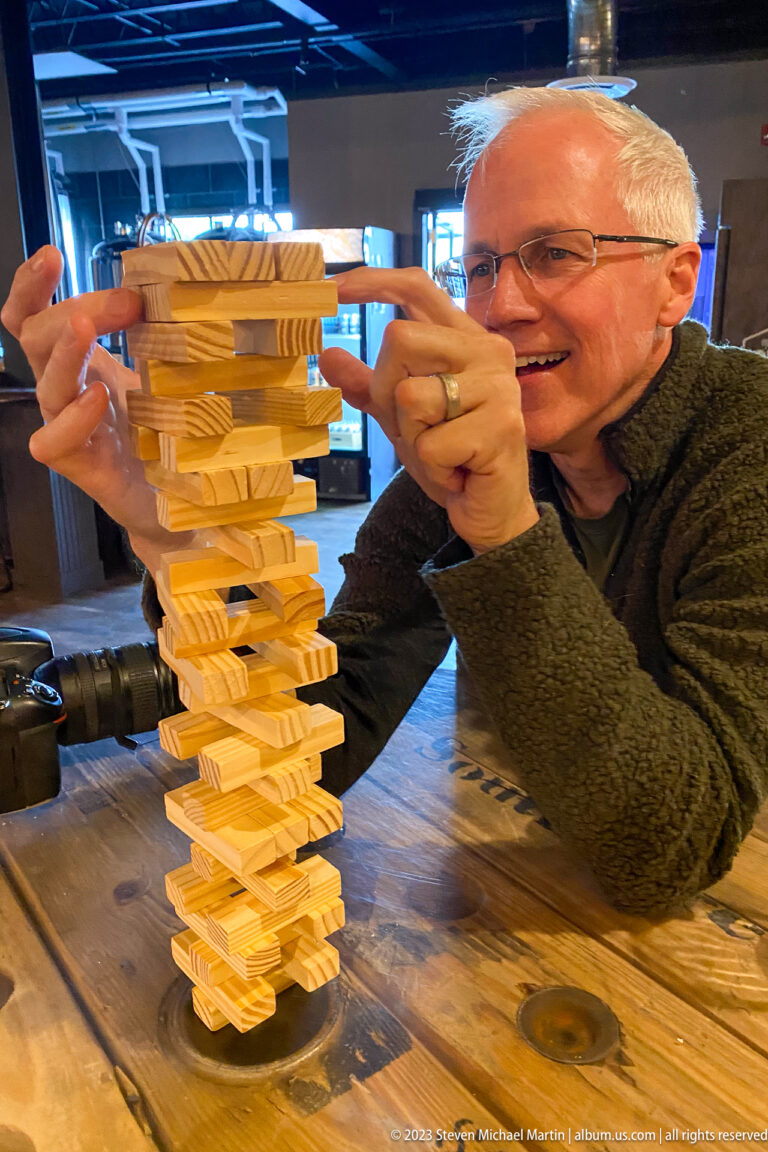 Man using both hands to adjust a block near the top of a tall, unstable wooden stacking tower.