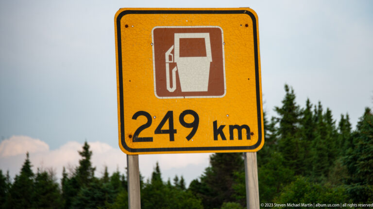 Yellow road sign with a gas-pump symbol and the distance “249 km” against conifer forest.