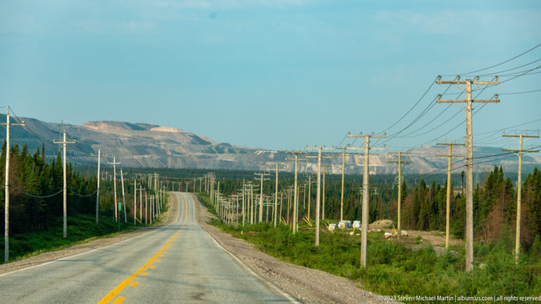 Straight paved highway with rows of utility poles leading toward a large terraced mine in the distance.