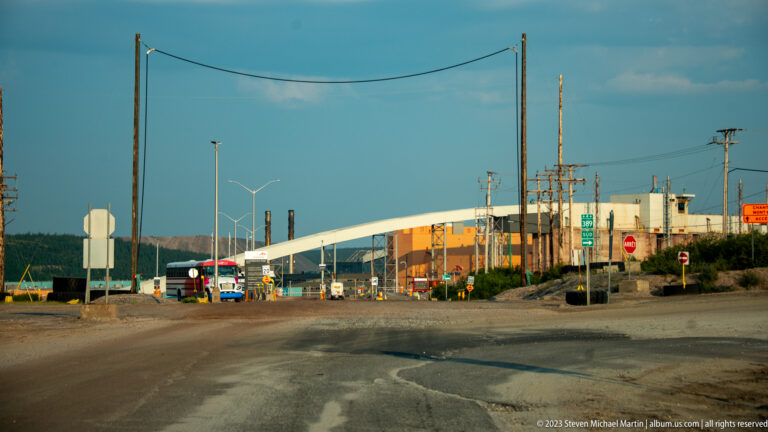 Industrial mine entrance with access controls, utility poles, large buildings, and an elevated enclosed conveyor.