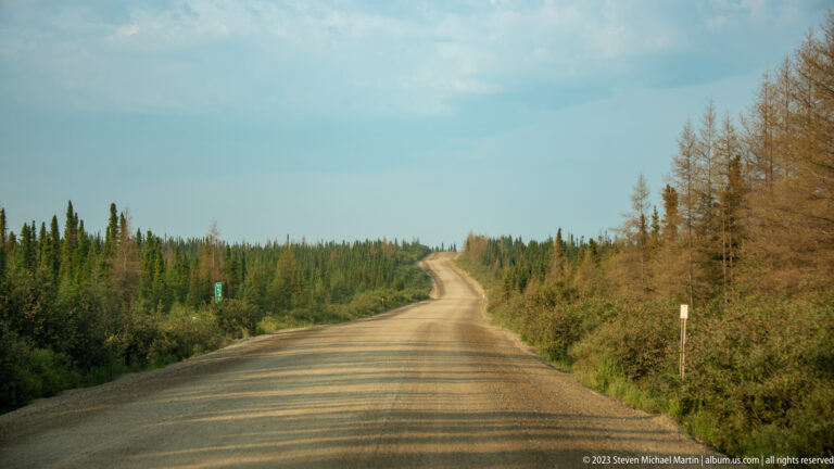 Long gravel road extending over low rises through dense boreal forest.