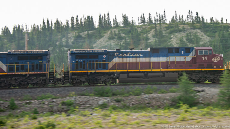 Two coupled blue, white, and red diesel-electric locomotives passing through rocky boreal terrain.