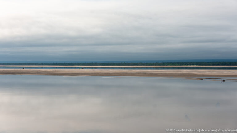 Still pale water and low exposed sandbars beneath a gray sky, with dark forest lining the far shore.