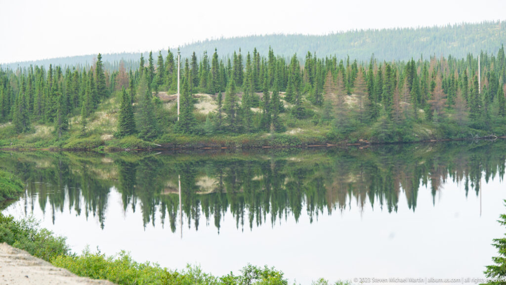 Still pond reflecting spruce trees and a low sandy forest bank.
