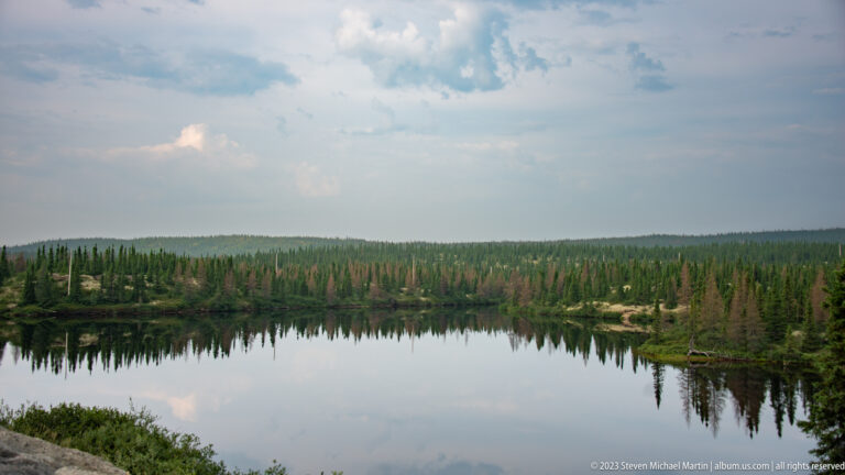 Wide calm lake reflecting a low conifer forest and cloudy sky.
