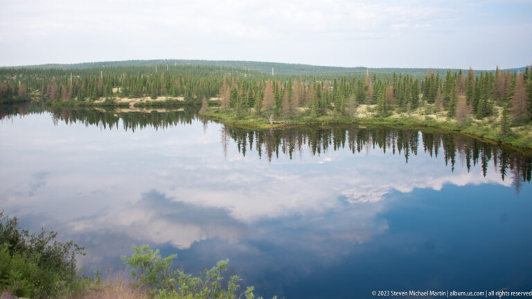 Calm boreal lake with near-perfect reflections of clouds and a low conifer shoreline.
