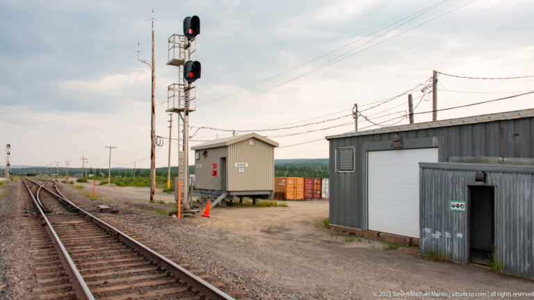 Railway tracks beside a signal mast, utility buildings, and overhead lines in a remote industrial corridor.