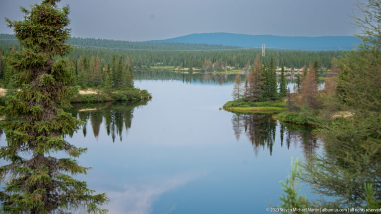 Calm boreal waterway with small wooded islets, conifers, and distant blue hills.