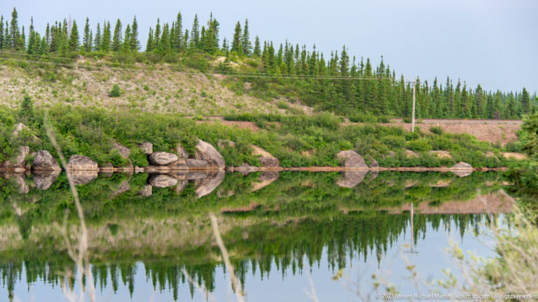 Still pond reflecting rounded shoreline boulders, shrubs, and sparse spruce on a pale rocky slope.