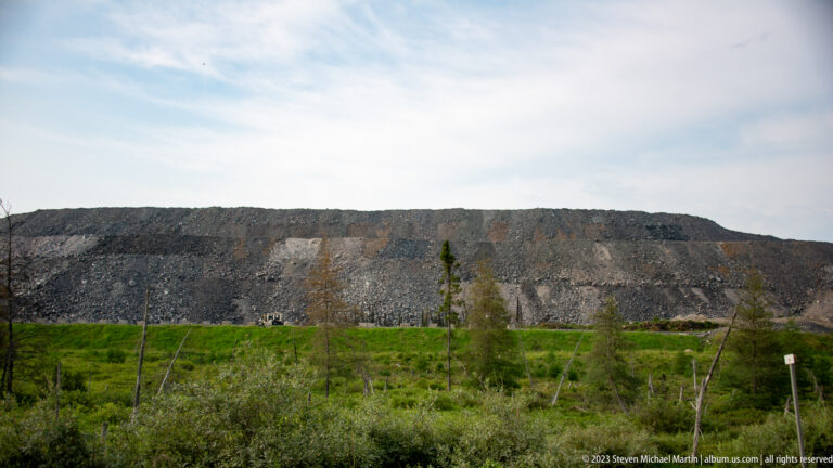 Long high embankment of broken gray rock rising behind shrubs and sparse boreal trees.