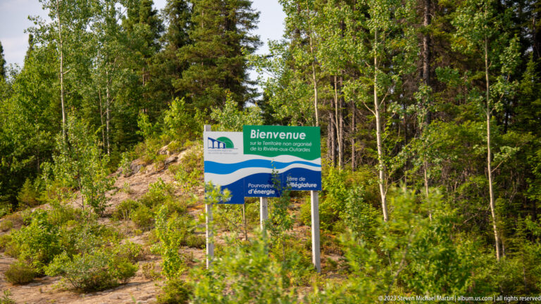 Green-and-blue French roadside welcome sign standing beside dense forest in Quebec.