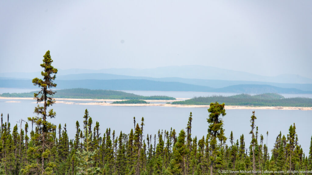 Elevated view over spruce forest to a broad reservoir with low islands and hazy layered ridges.