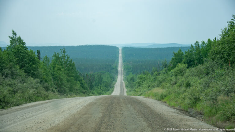 Long straight gravel road crossing repeated hills through dense boreal forest toward distant blue ridges.