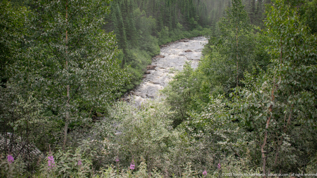 Rocky, fast-flowing river seen through dense green boreal forest from an elevated viewpoint.