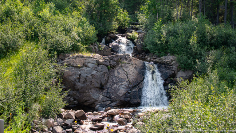 Two-tier waterfall dropping over dark bedrock amid dense green forest and scattered boulders.