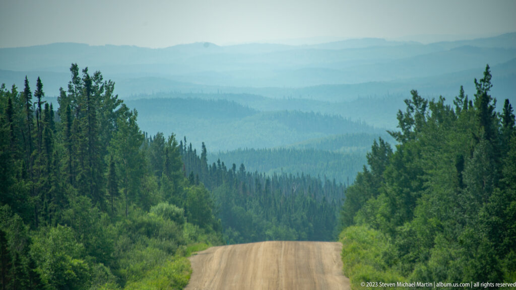 Gravel road descending through boreal forest toward many hazy, layered hills in the distance.
