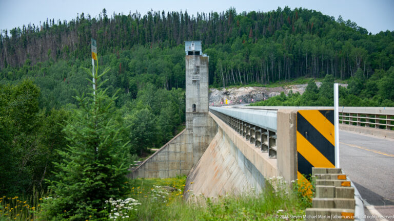 Roadway crossing the crest of a large concrete dam beside a tall rectangular service tower and forested slopes.