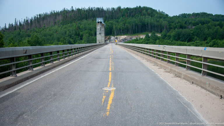Centered view along a two-lane roadway crossing a dam, with railings and a tall concrete tower ahead.
