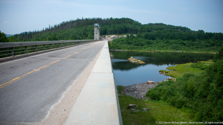 Dam-top roadway beside calm reservoir water, a small rocky islet, and green shoreline vegetation.