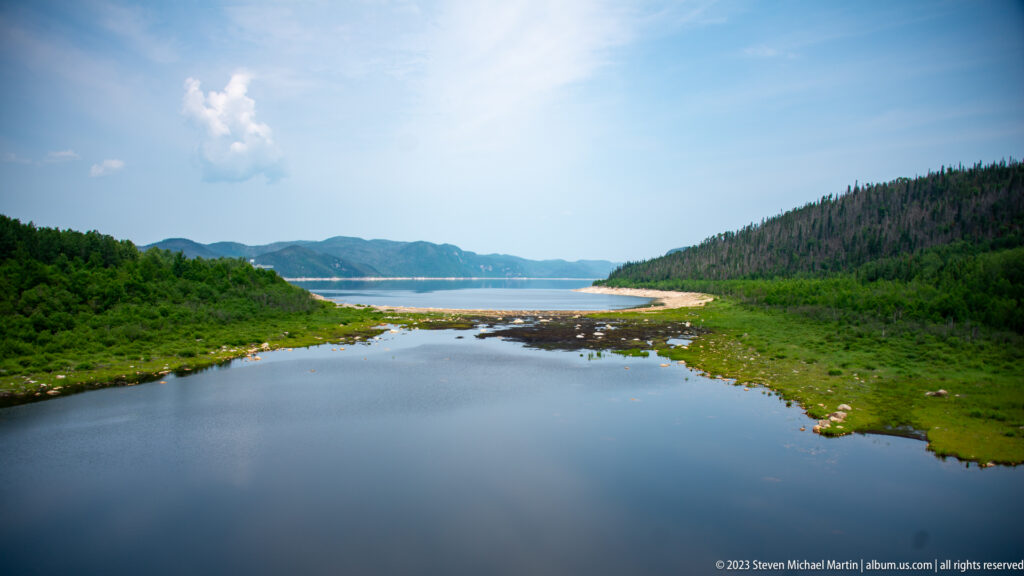 Calm reservoir inlet with marshy green banks, exposed rocks, forested hills, and open water beyond.