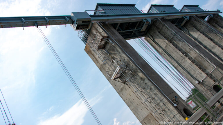 Low-angle view up a tall concrete dam tower with ladders, platforms, heavy cables, and mechanical equipment.