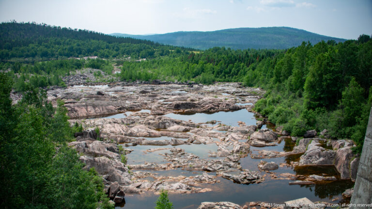 Wide exposed bedrock river channel with scattered pools, surrounded by dense forest and a low hill.