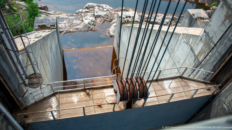 Overhead view of heavy hoisting cables, large sheaves, railings, and a concrete dam gate bay.
