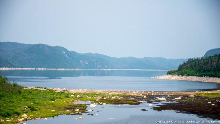 Wide reservoir between forested hills, with marshy pools and exposed stones in the foreground.
