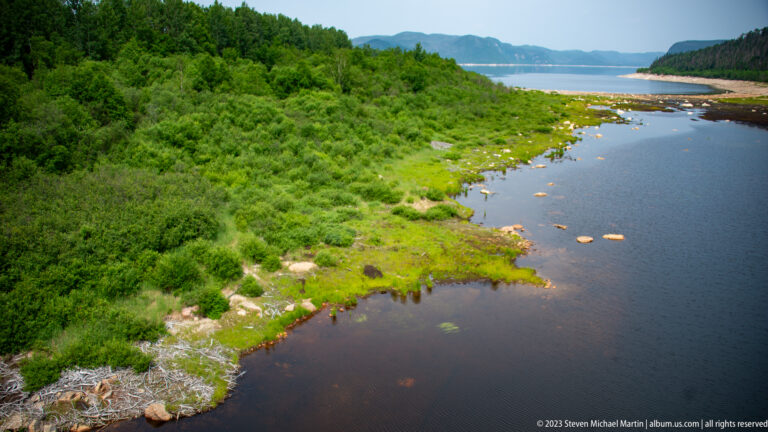 Dark narrow inlet bordered by lush shrubs and rocks, opening toward a distant reservoir between wooded hills.